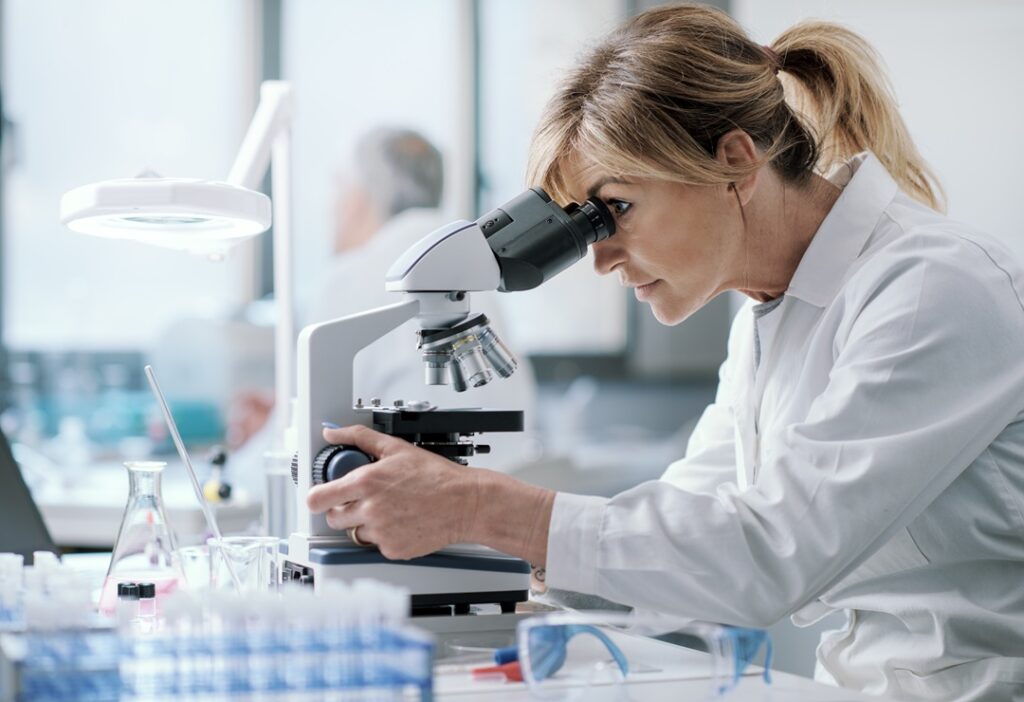 Kosmetikstudium, eine blonde Frau sitzt im Labor und schaut durch ein Mikroskop